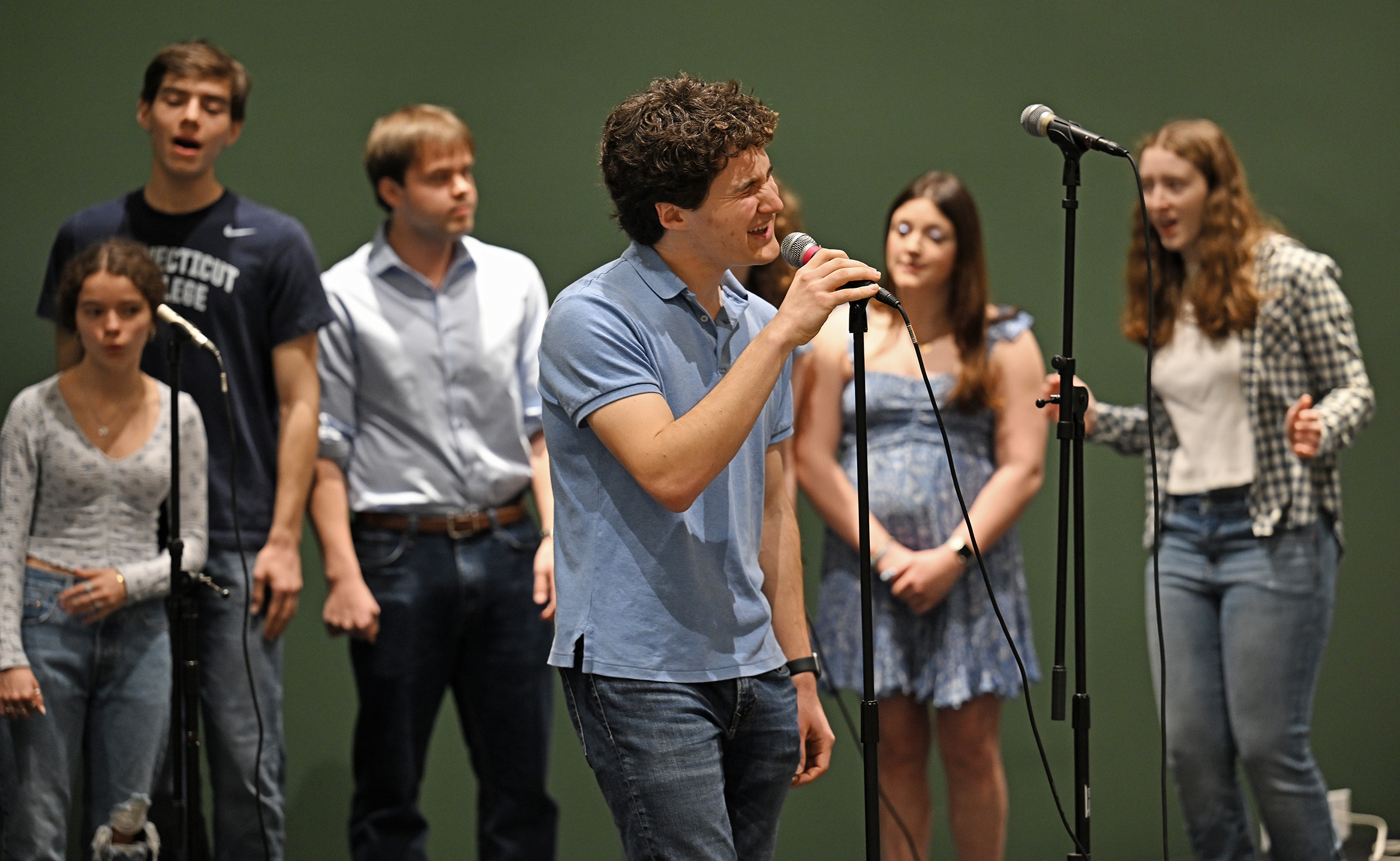 A male a cappella singer solos a a microphone with the full a cappella group behind him singing.