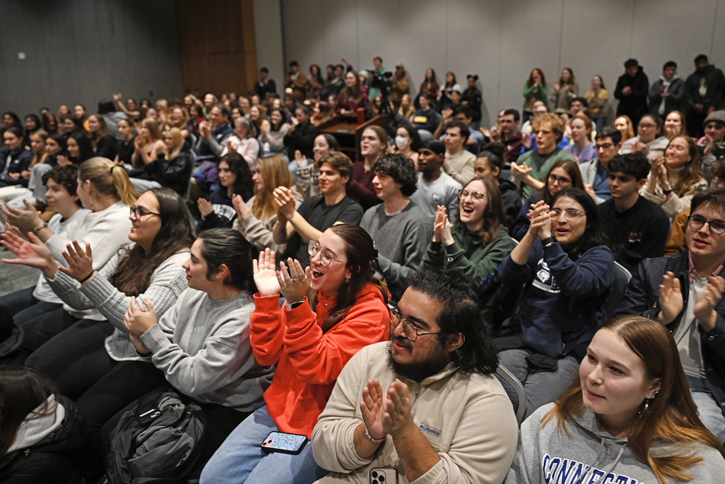 A large audience of college students sit and applaud at an a cappella show.