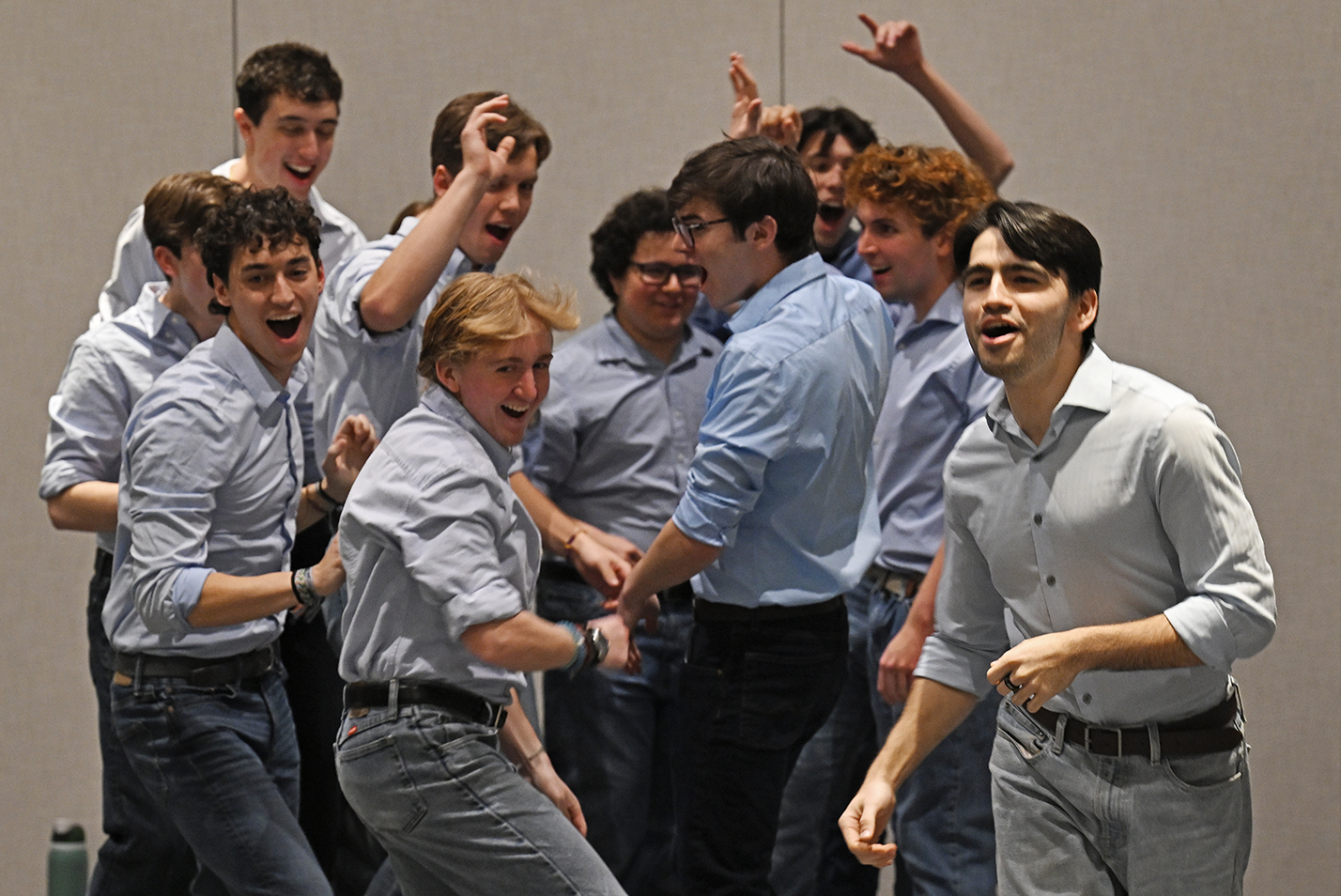 A group of male a cappella singers in blue shirts and ties shout a cheer as they turn to run onto the stage.