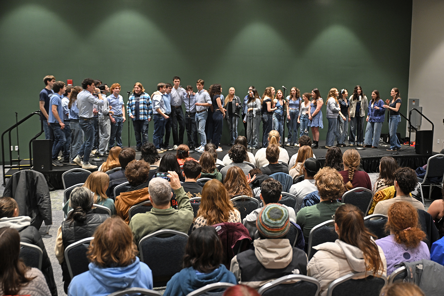 A large group of college a cappella singers perform on a ballroom stage.