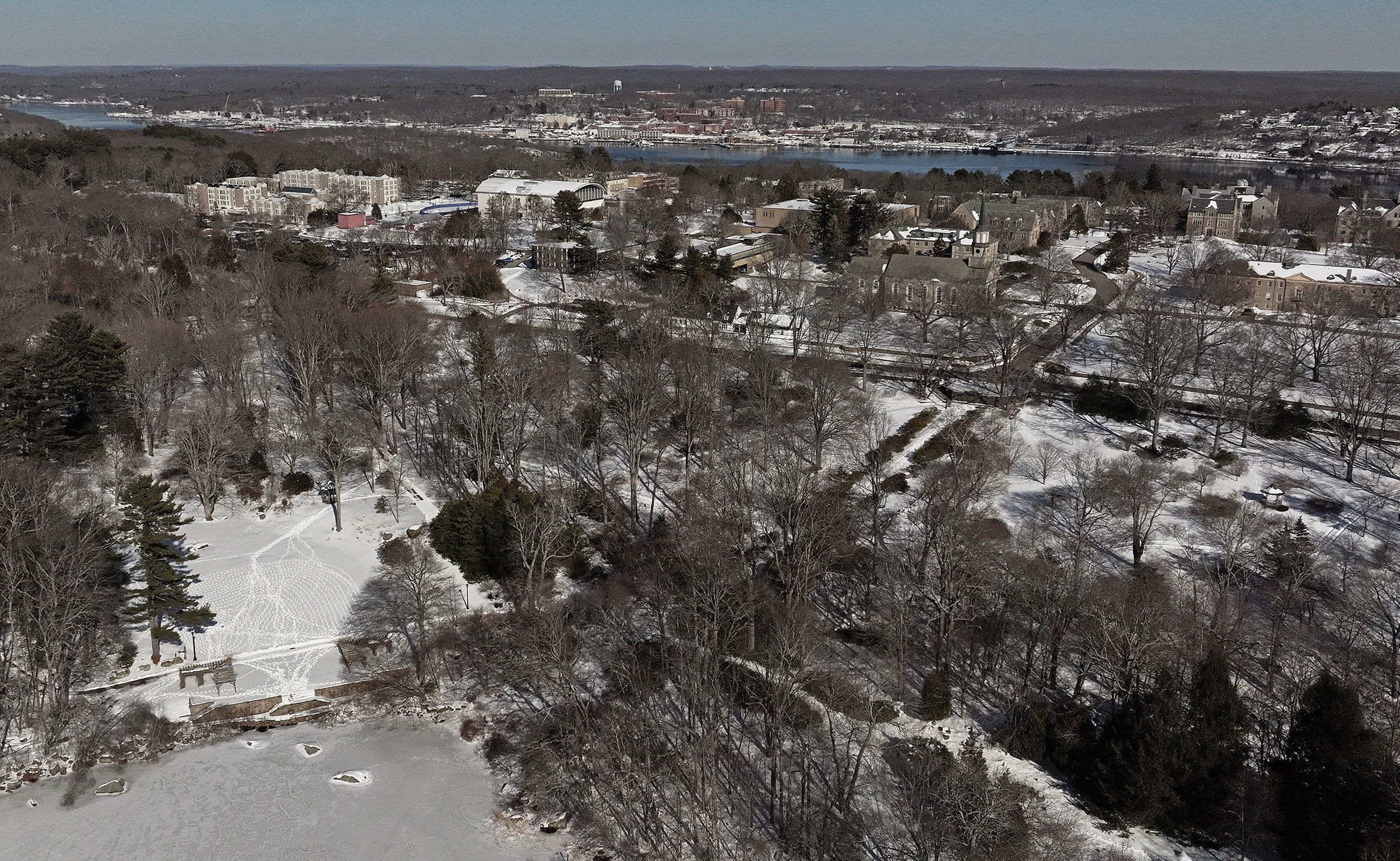 Aerial view of a snow-covered wooded area and frozen pond with a small college campus in the distance.