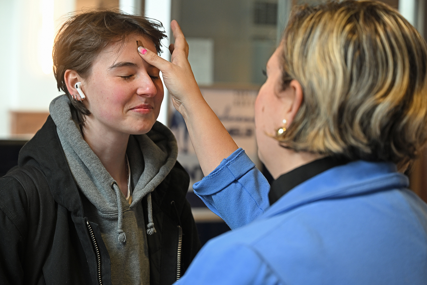 A college chaplain in blue blazer imparts ashes on the forehead of a student with short curly hair and wearing a scarf.