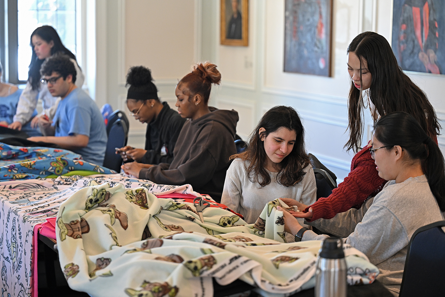 Students gathered around rows of tables work with fleece material to make blankets.