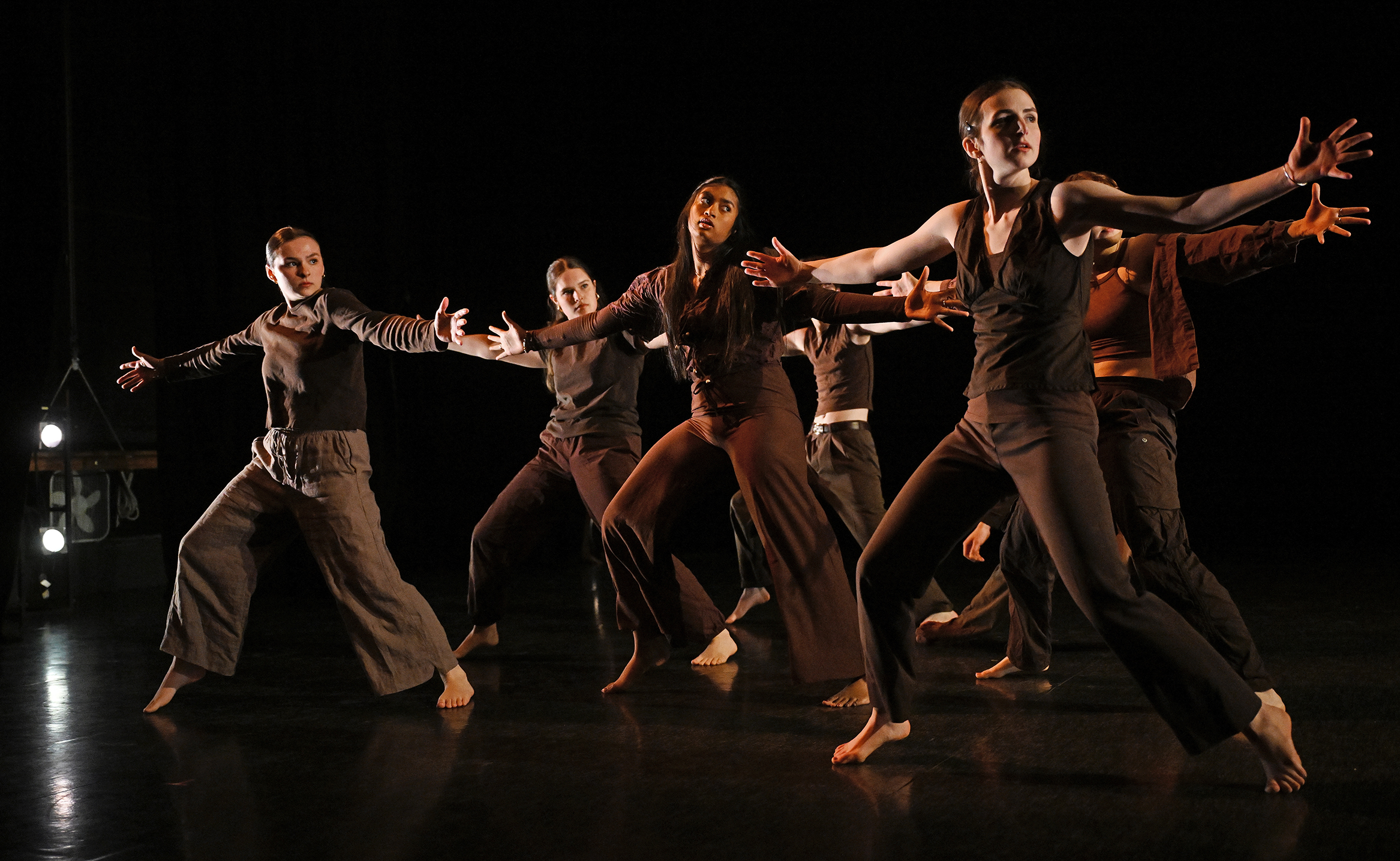 Student dancers in brown tops and pants with arms outstretched perform in a college dance studio.