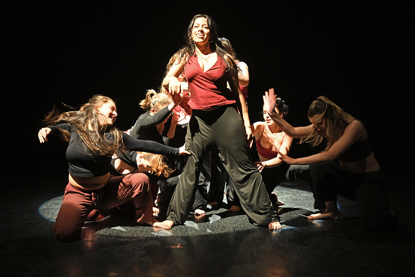 Student dancers in a spotlight dance on their knees around one standing dancer in a college dance studio.