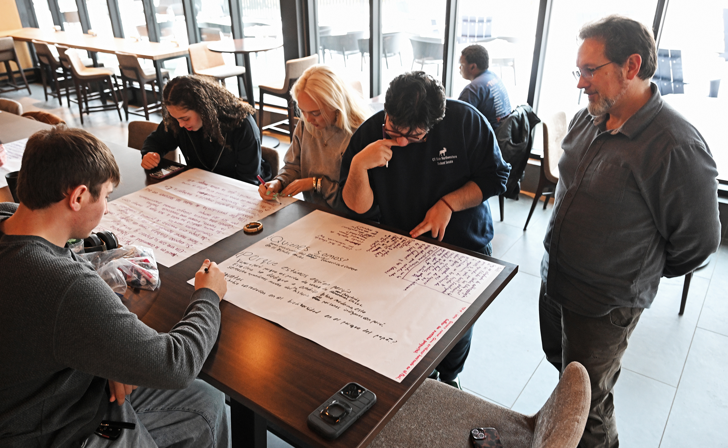 A college professor meets with students working a large sheets over paper on a student pub table.