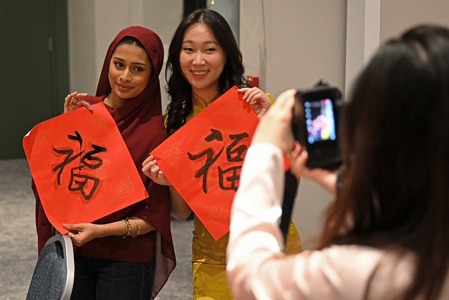 Two female students, one with a red headscarf, hold up Asian calligraphy examples for a photographer.