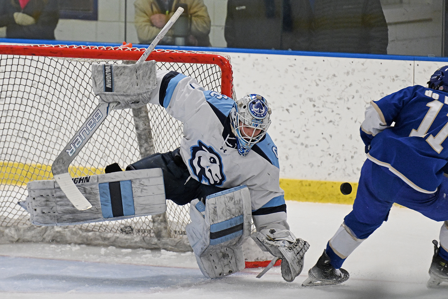 A men's hockey goalie leans to the right partially leaving the ice to block the puck.