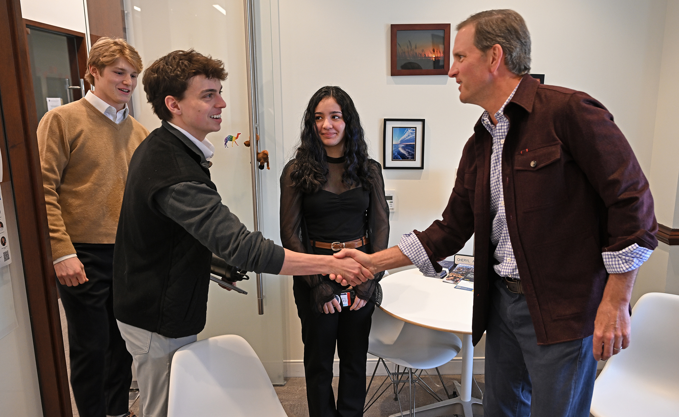 A businessman in casual dress shakes hands with a college student in jacket and tie as two other students look on in a college office.