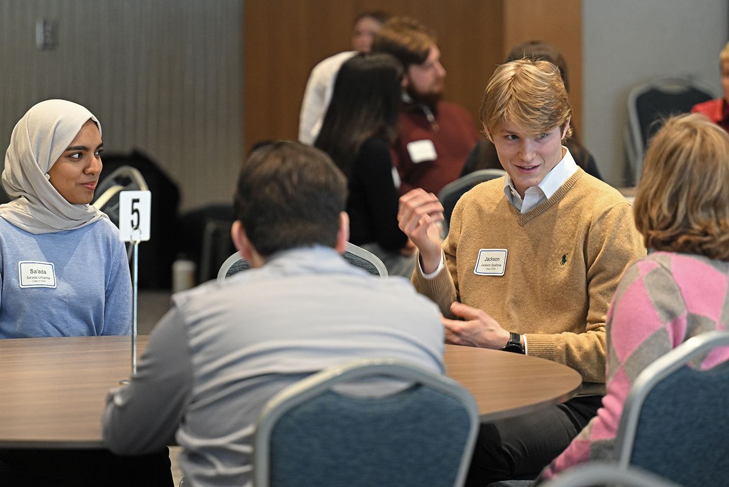 A male college student in a yellow sweater makes a point while seated around a table with other students.