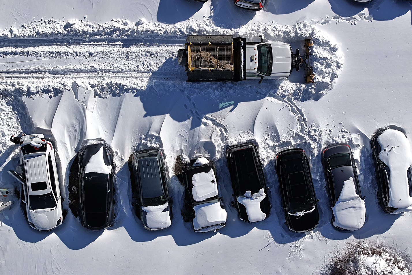 Seen from overhead, a plow truck pushes snow in a parking lot covered by a blizzard.