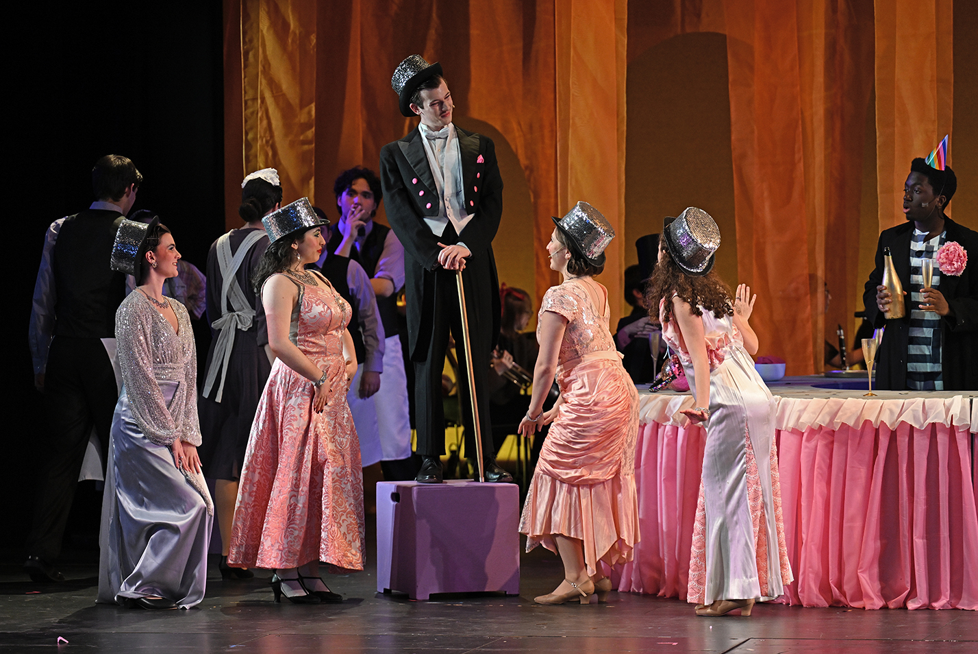 Students in fancy costumes, one in suit and top hat standing on a platform, act on stage.