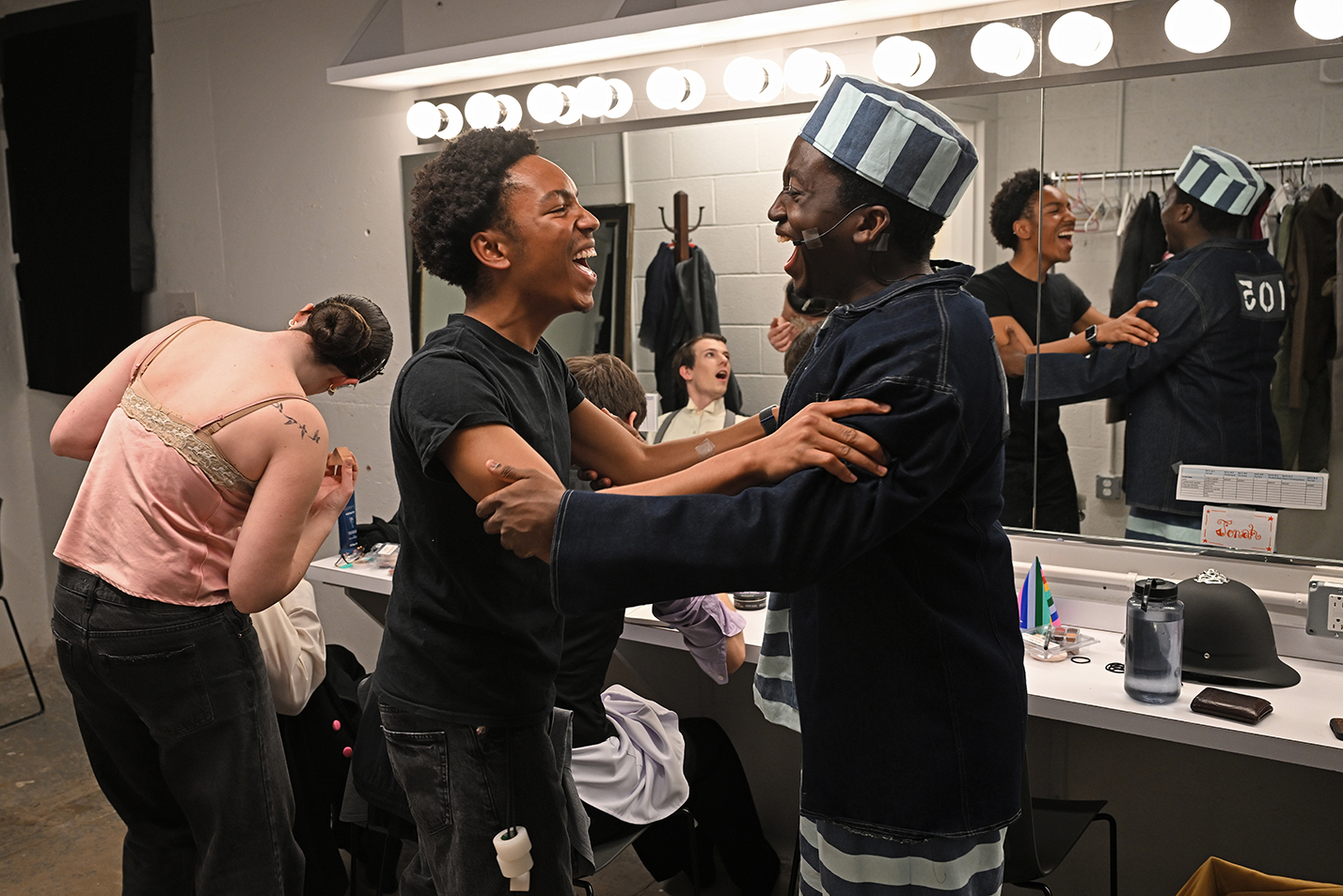 A stage technician and an actor in prison garb embrace and cheer as they get primed for a musical theater production in a dressing room.