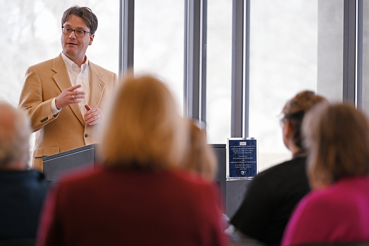 A man in a tan sport coat stands at a podium and makes remarks before presenting an award, on the table to his left, at a college library.