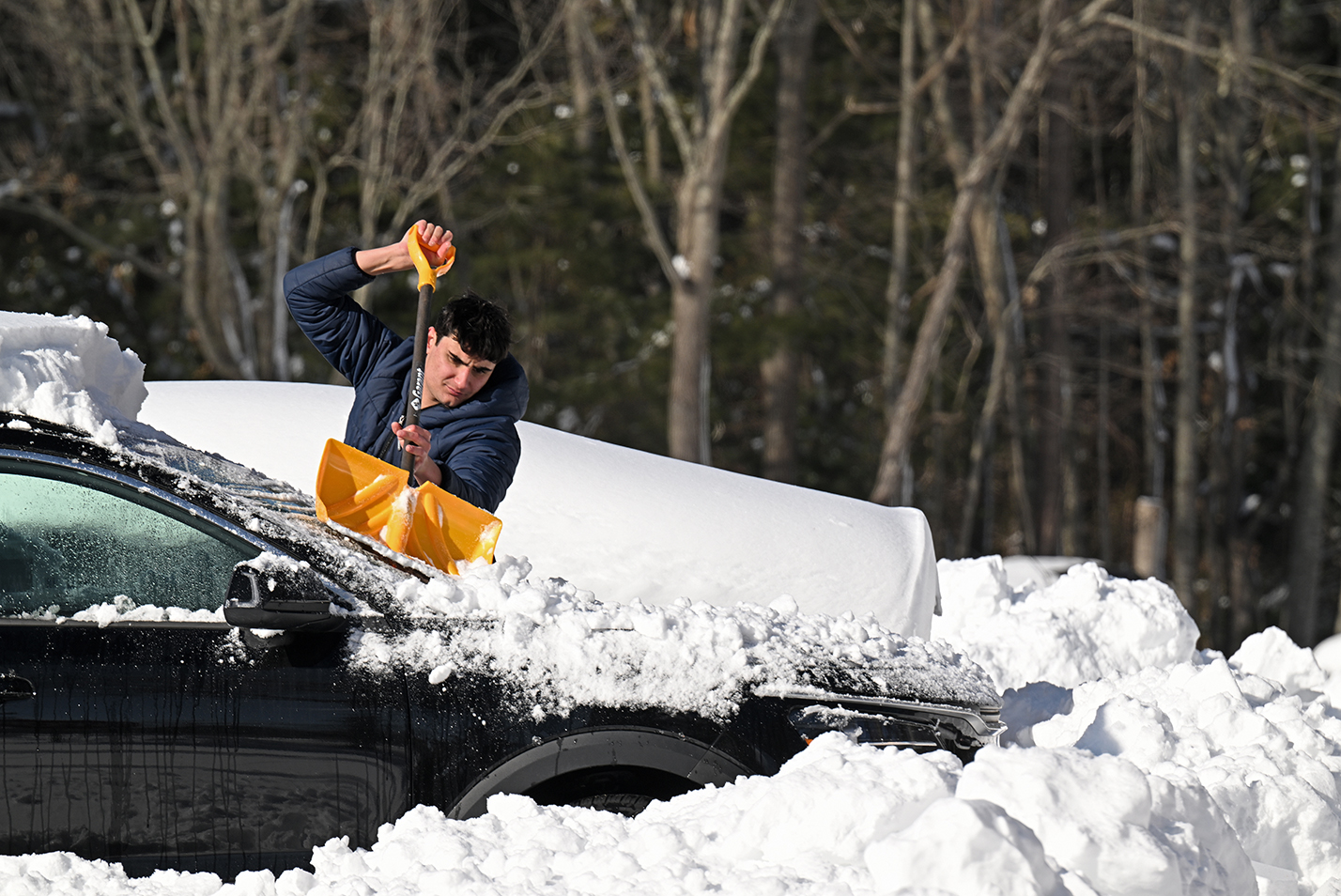 A college student uses a snow shovel to clear snow off his car.
