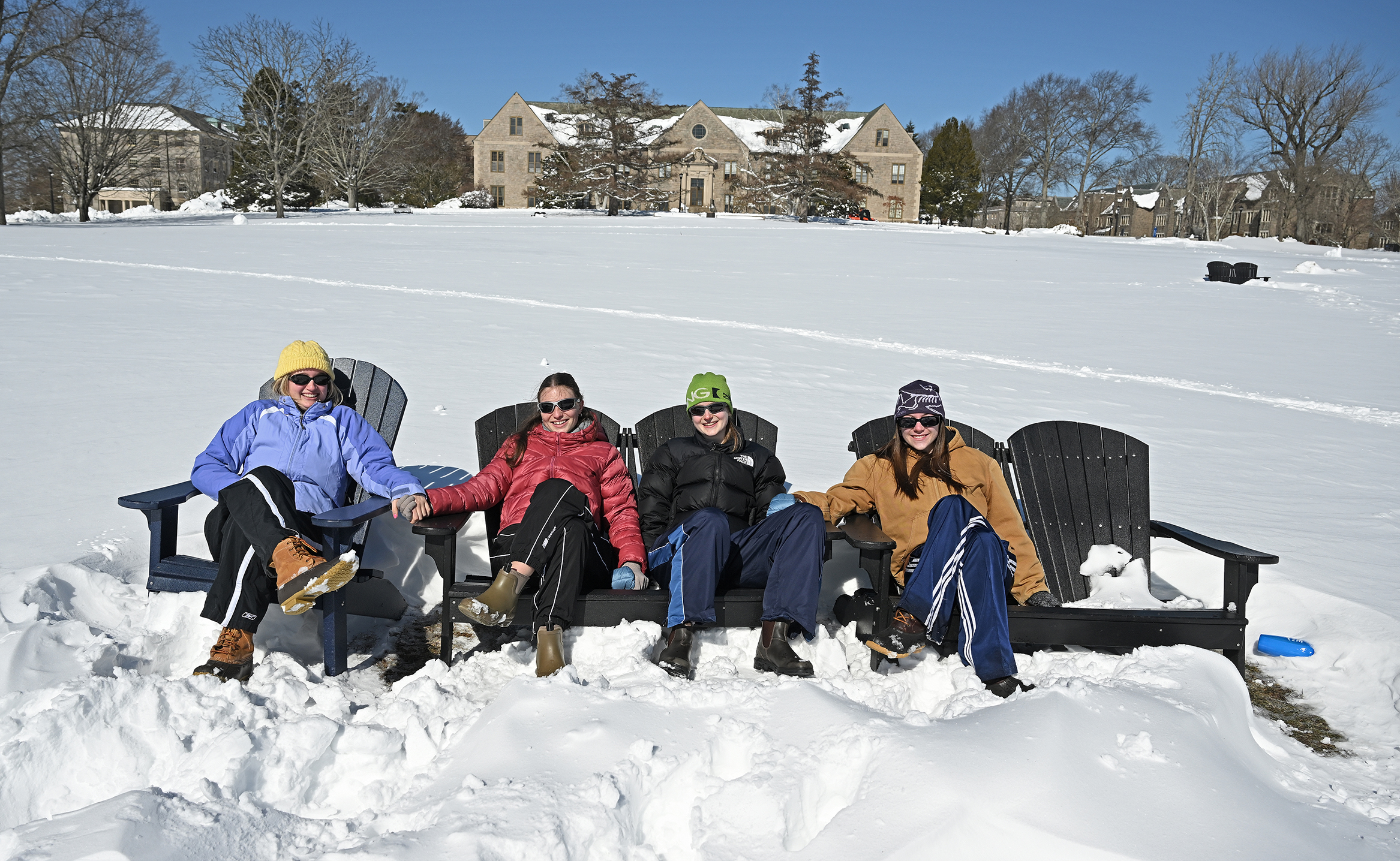 Four female college students in winter coats sit in Adirondack chairs in the middle of a large snowy field.