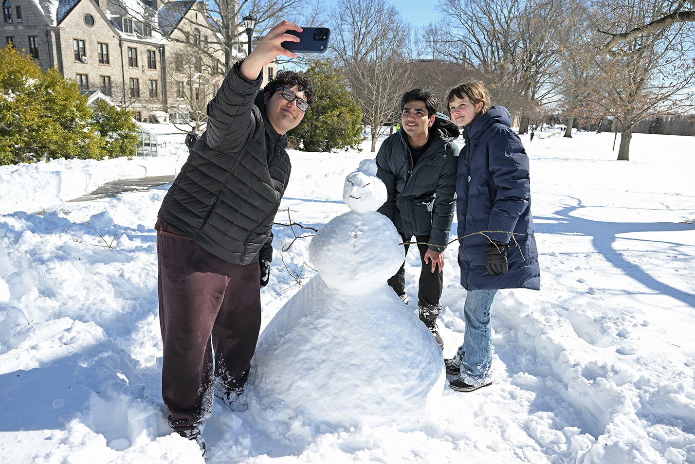 Three college students gather around a snow person they built on the edge of a college green and take a photo together.