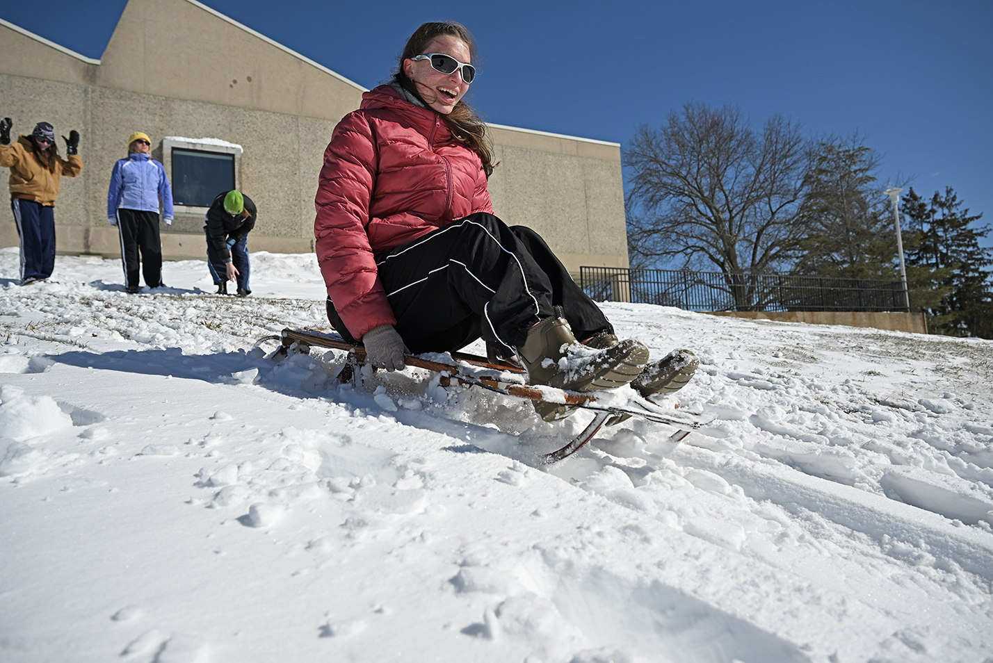 A college student in a red puffy winter coat slides down a snow-covered hill on a metal rail sled.