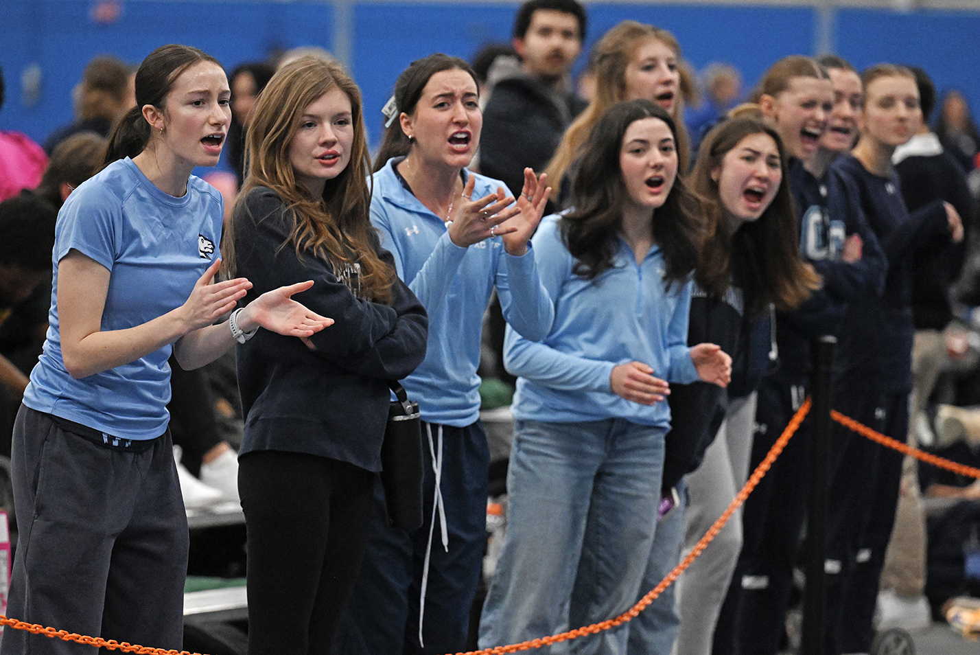 A group of female track and field athletes, some in warm ups, some in street clothes, gather on the side of the track to cheer on a teammate.
