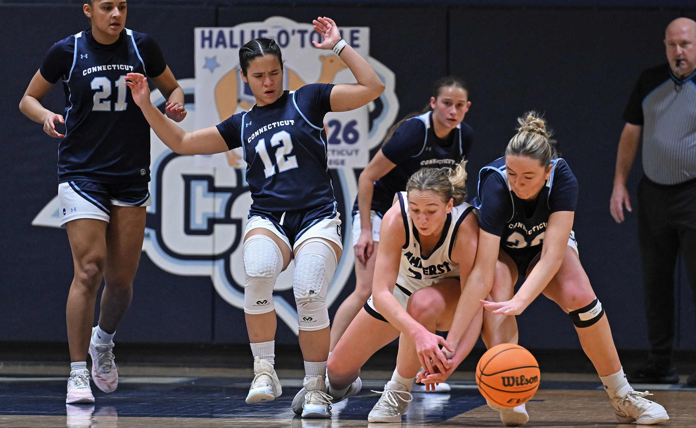 Two women's basketball players scramble for a loose ball at the rights side of the frame as three teammates scramble after the play.