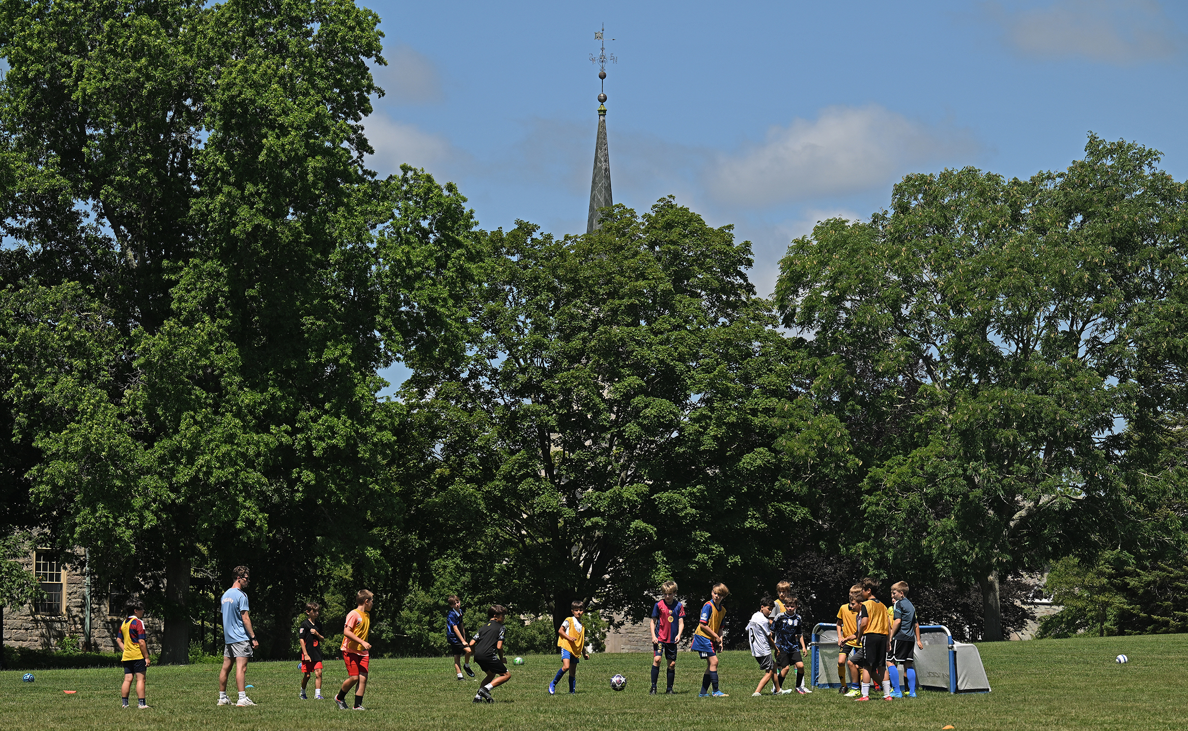 Youth soccer players in colorful shirts play on a college green with a row of trees and a chapel steeple looming in the background.