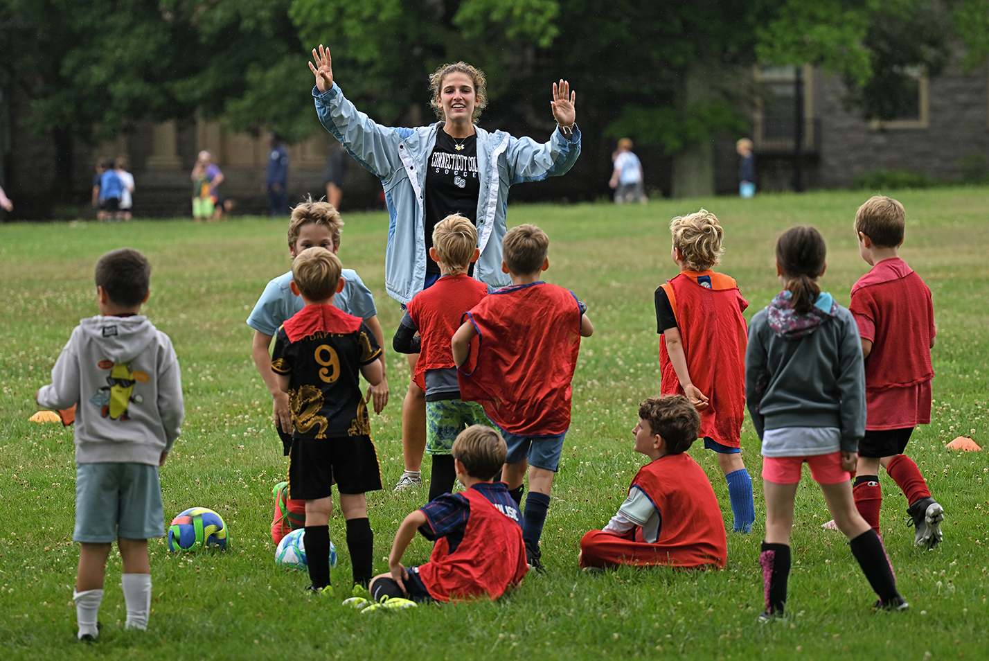 A female college soccer player in a light blue windbreaker and a college soccer t-shirt raises her arms to give direction to a group of youth soccer players wearing red shirts on an open college green.