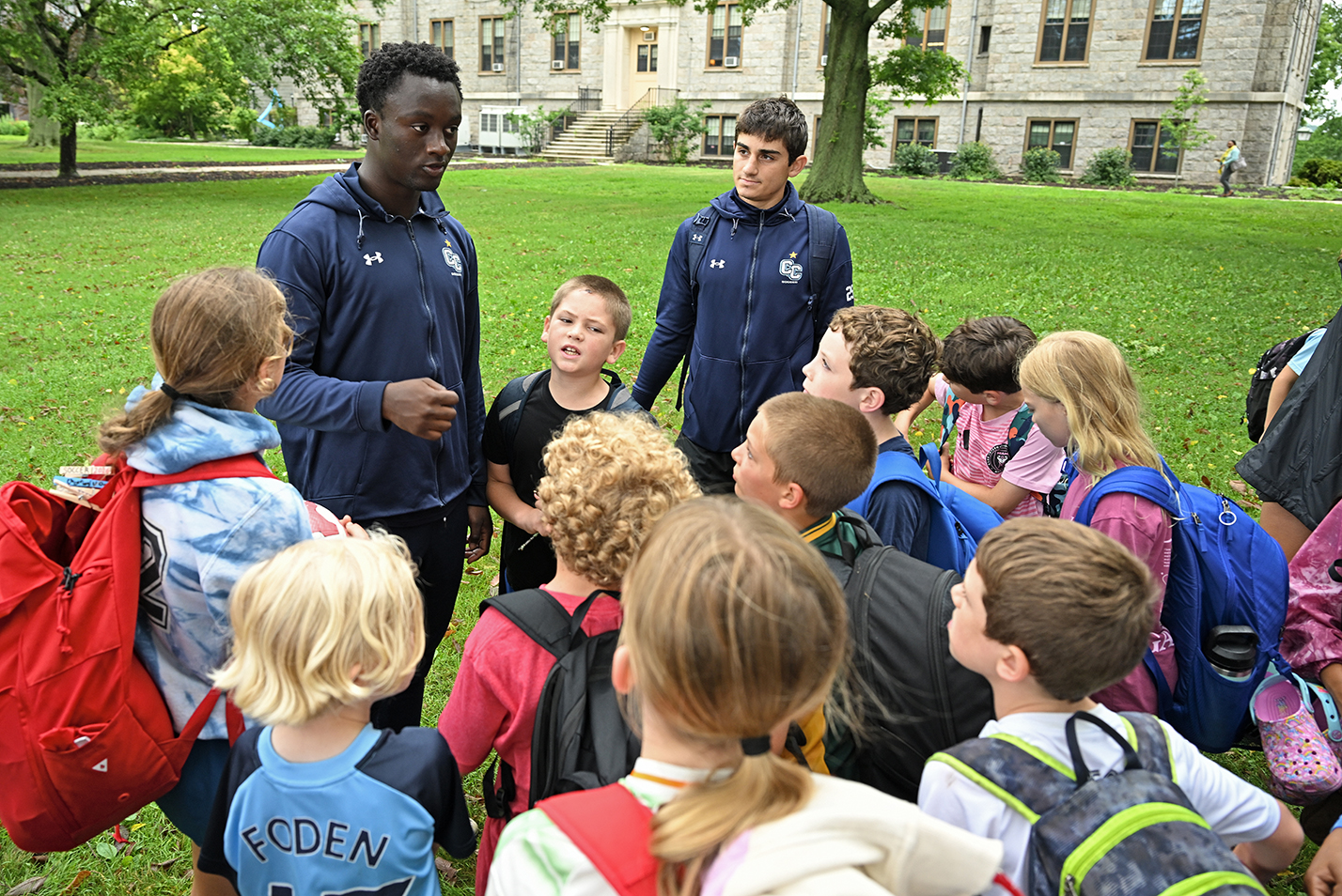 A pair of men's college soccer players in dark blue zipper hoodies with a CC logo speak to a group of youth soccer players on a wooded green outside a granite academic building.