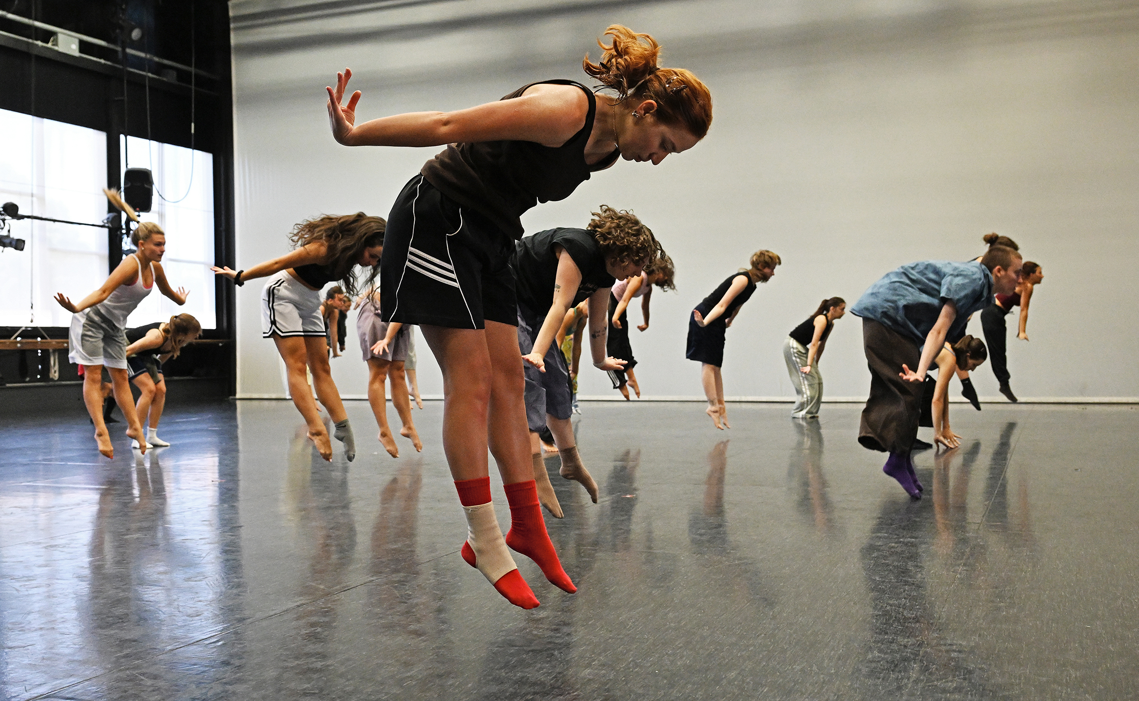 Dance students spring off the floor with pointed toes while also bent over forward with their hands thrown out to the sides in a dance studio space.