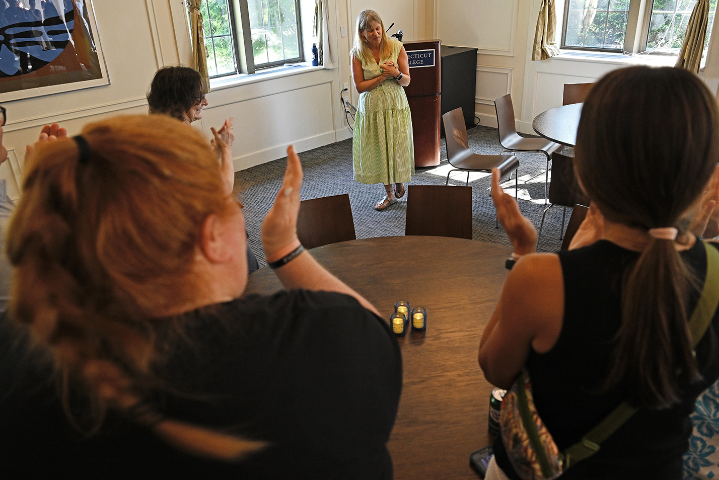 A female college professor in a light green vertical stripe dress clasps her hands to her chest as a crowd stands and applauds around her.