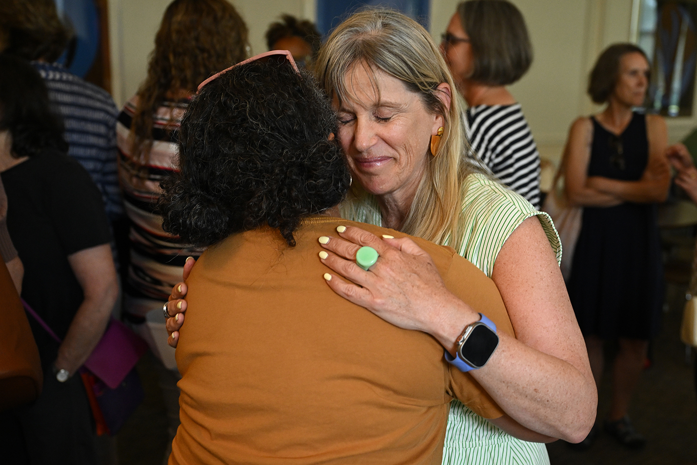 A female college professor in a light green vertical stripe dress embraces a colleague in a brown shirt amidst a crowd of colleagues.