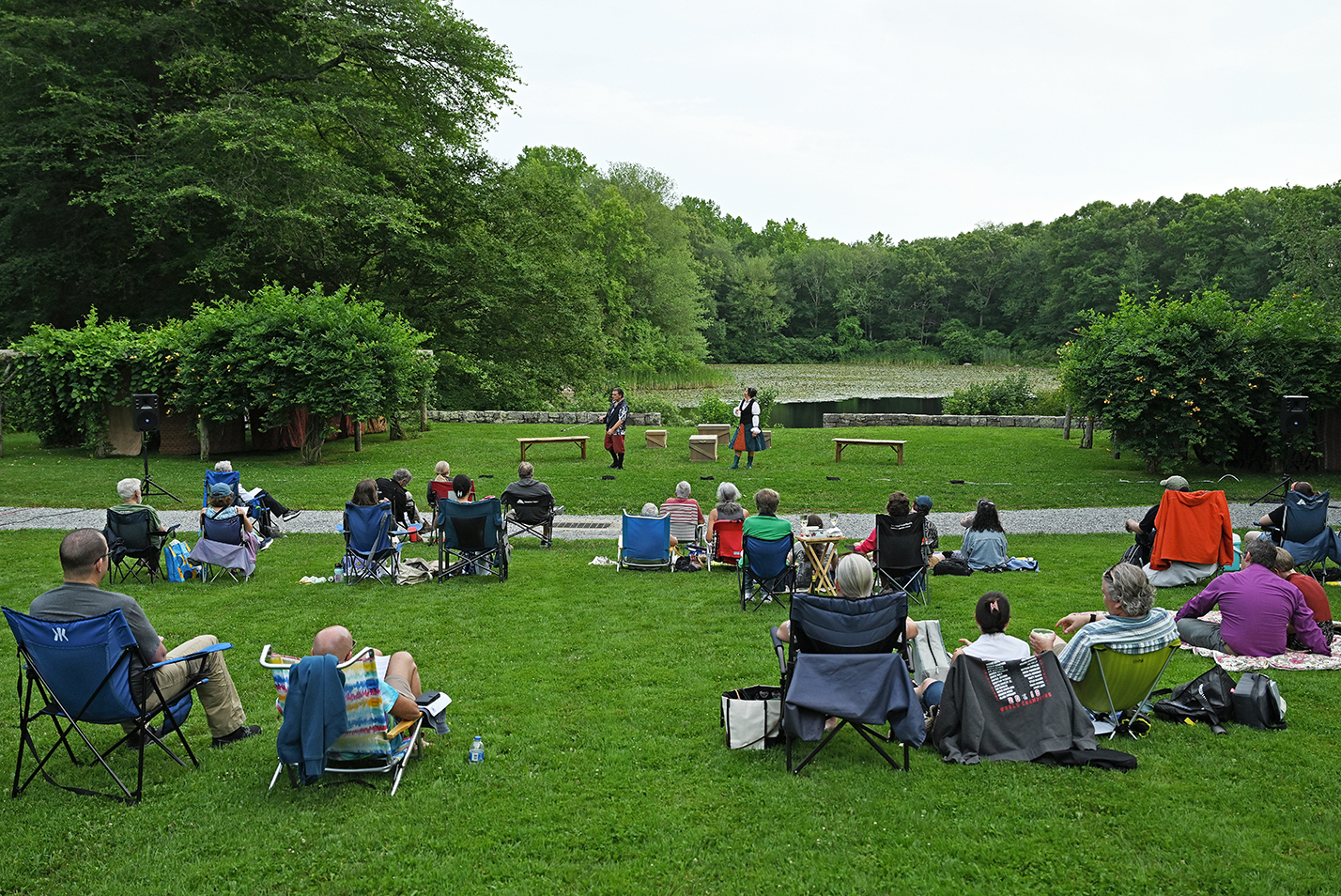 An audience seated in folding chairs and on blankets on the grass watch a performance of a Shakespeare play in an arboretum amphitheater with a pond in the background.