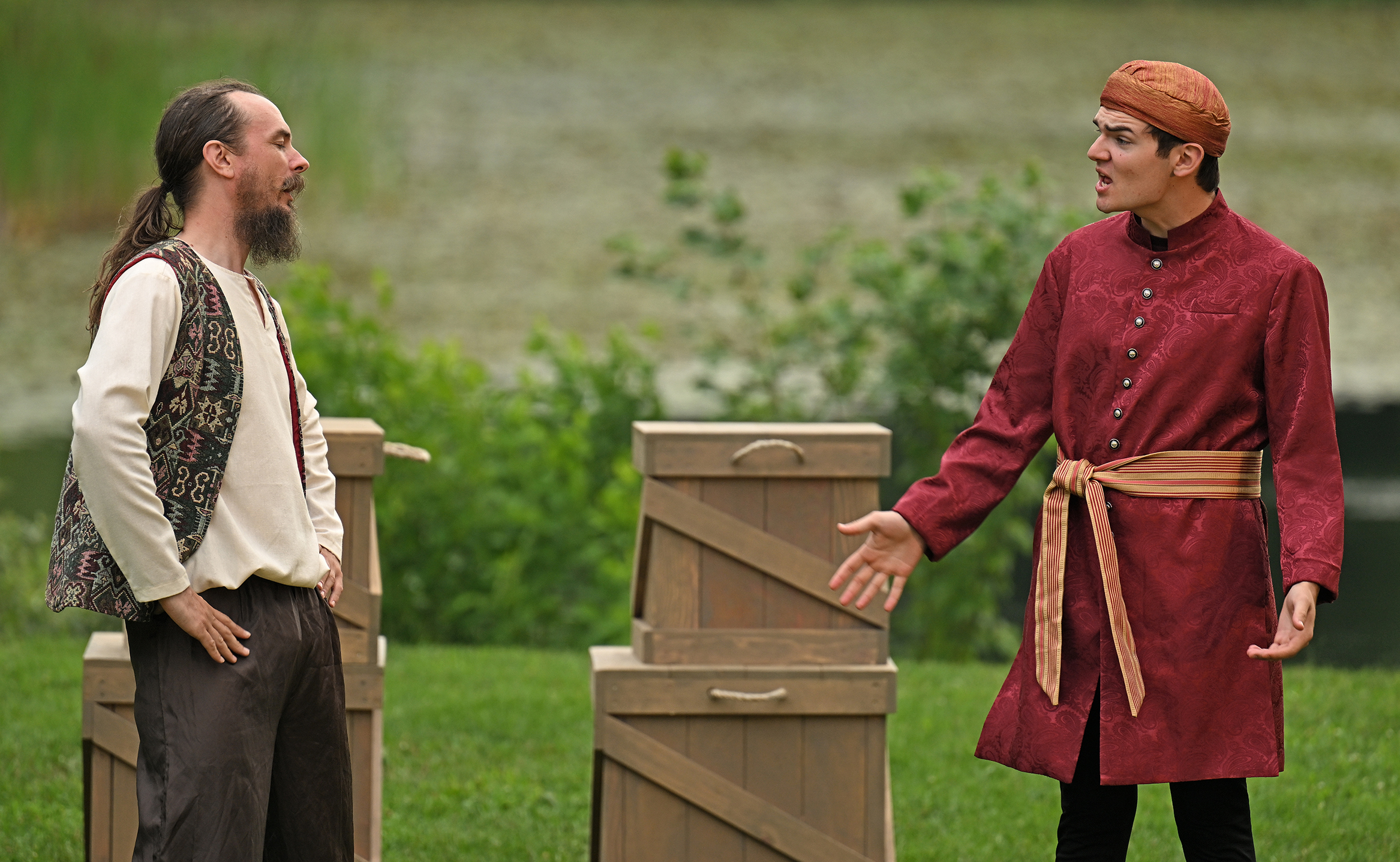 A pair of Shakespearean actors, one dressed as a sailor, one in a red jacket with orange sash belt and skullcap perform on an outdoor stage with prop boxes in the background.
