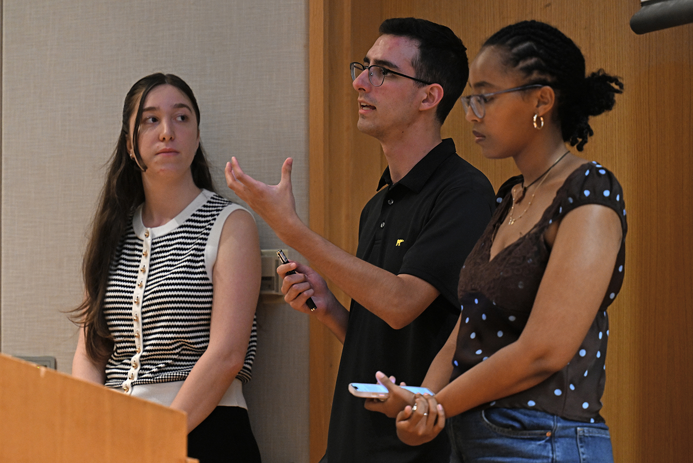 A male college student in dark polo shirt and glasses gestures while speaking standing between two female college students at a podium.