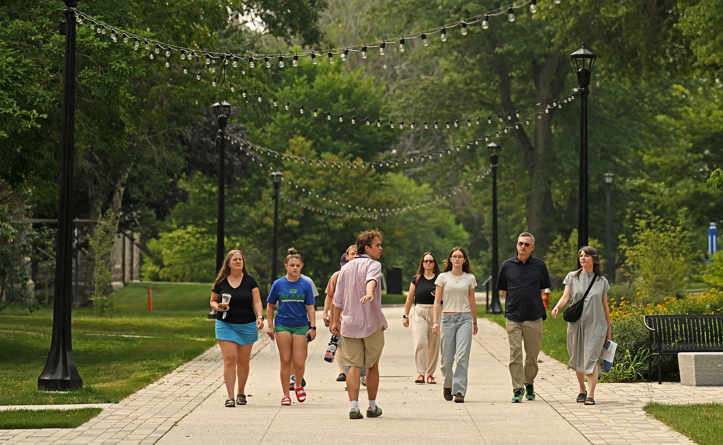 A male college student in baggy shorts and an untucked long-sleeve shirt, with the sleeves rolled up, walks backwards and gestures while leading a tour along a pedestrian walkway lined with tall black lampposts with little accent lights strung diagonally overhead.