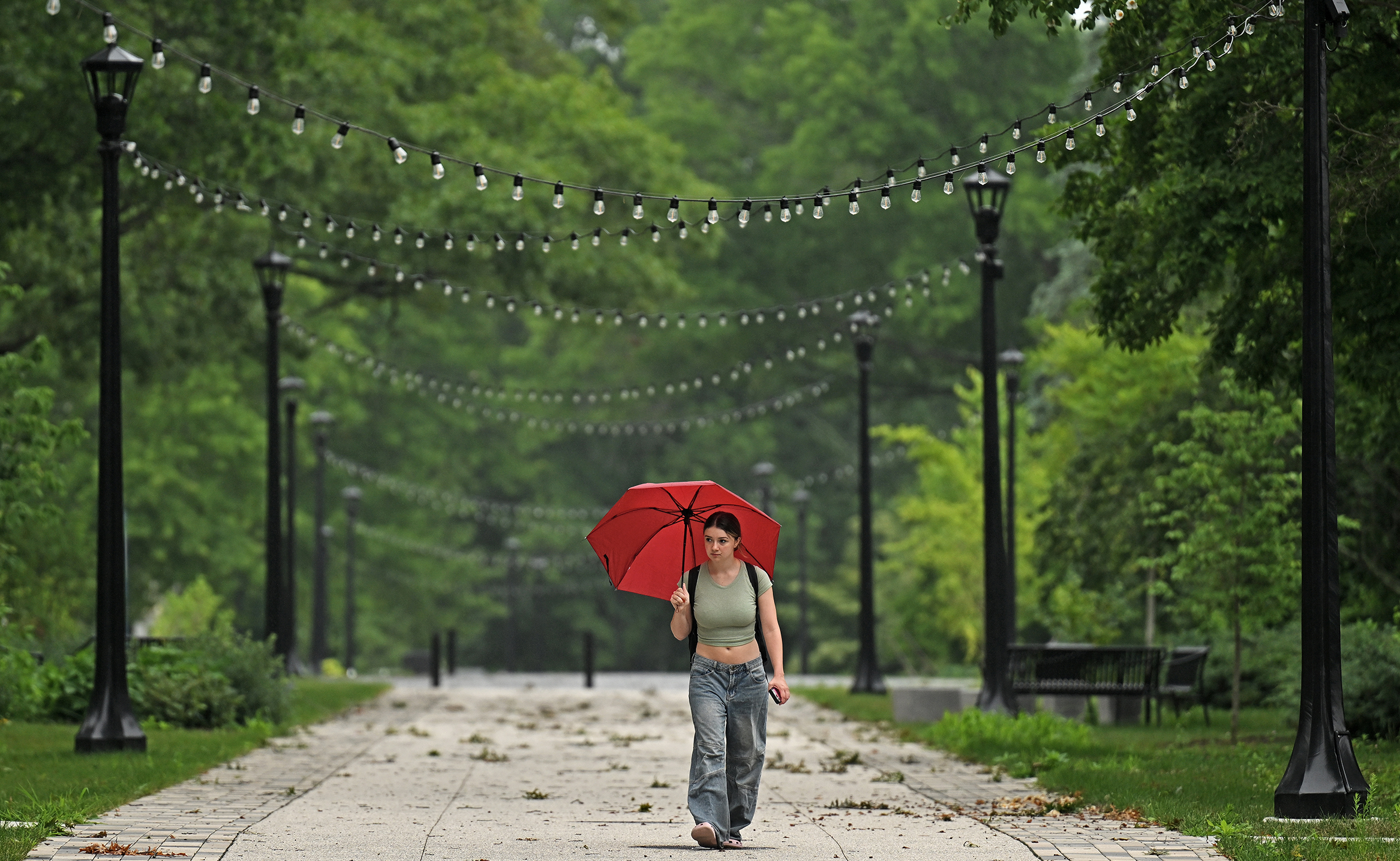 A female college student in crop top and jeans carries a bright red umbrella walking along a pedestrian walkway with small lights strung overhead from the lampposts and scattered leaf debris on the ground.