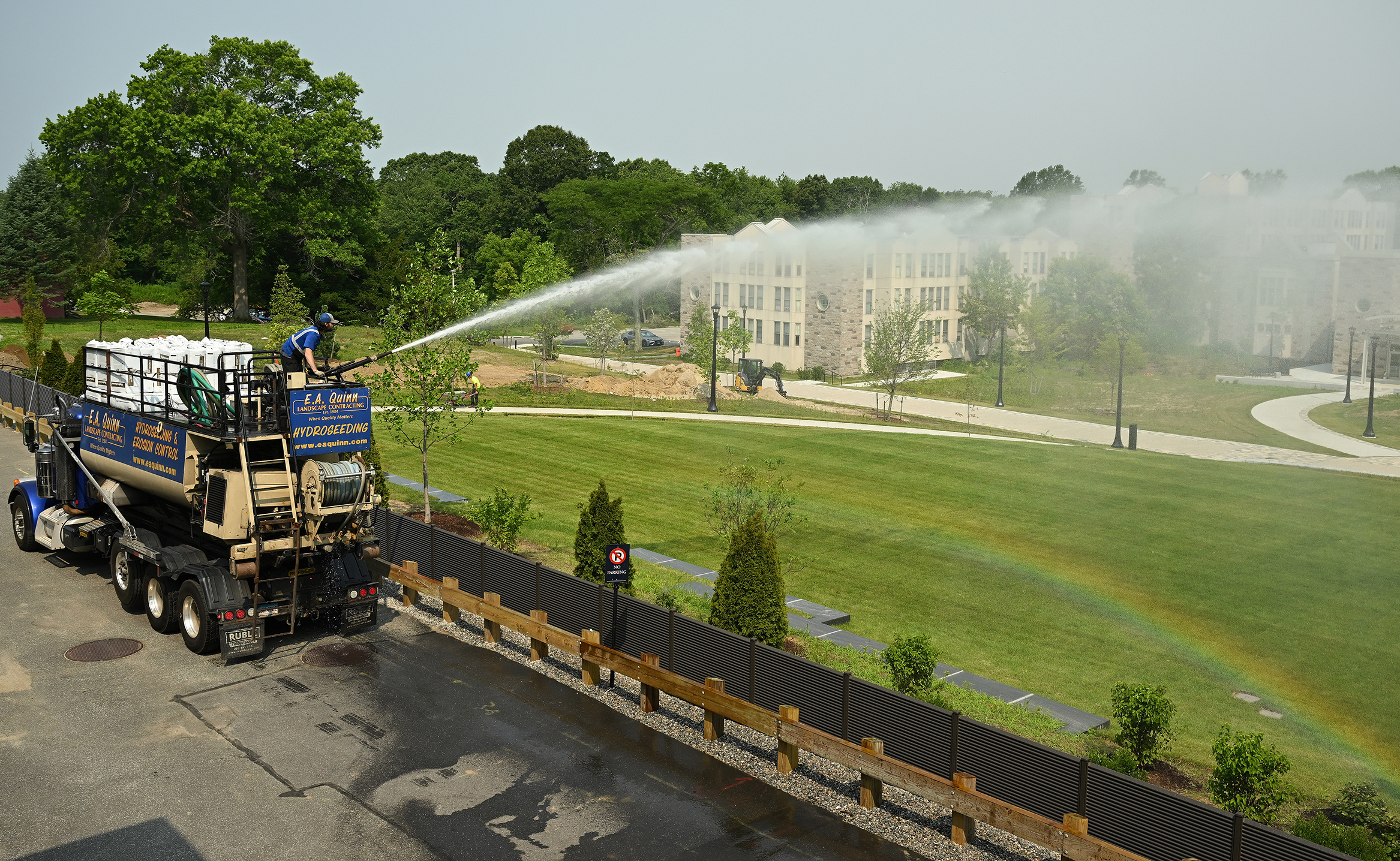 A landscaping crew with a large tanker truck sprays water over a newly planted sod field creating a rainbow under the stream of water.