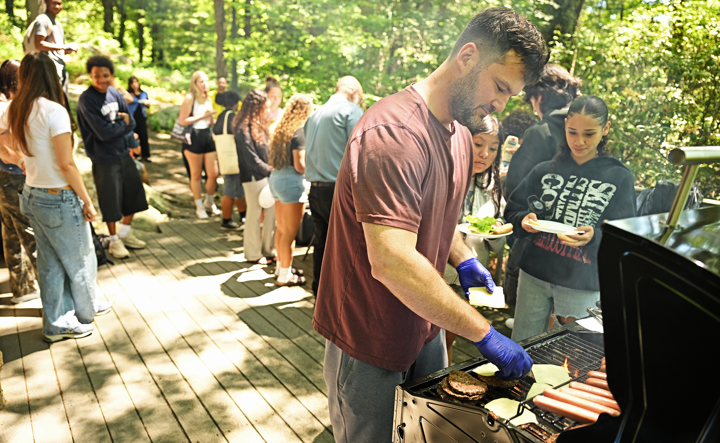 A man wearing blue food-service gloves places slices of cheese on hamburger patties cooking on a grill on an outdoor deck surrounded by trees as high school students wait wit plates in hand.