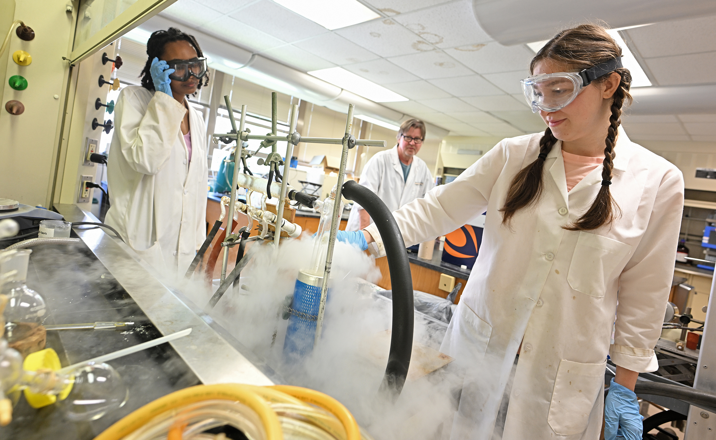 A college professor watches two students in white lab coats as fog from liquid nitrogen bubbles up from a flask in a college chemistry lab.