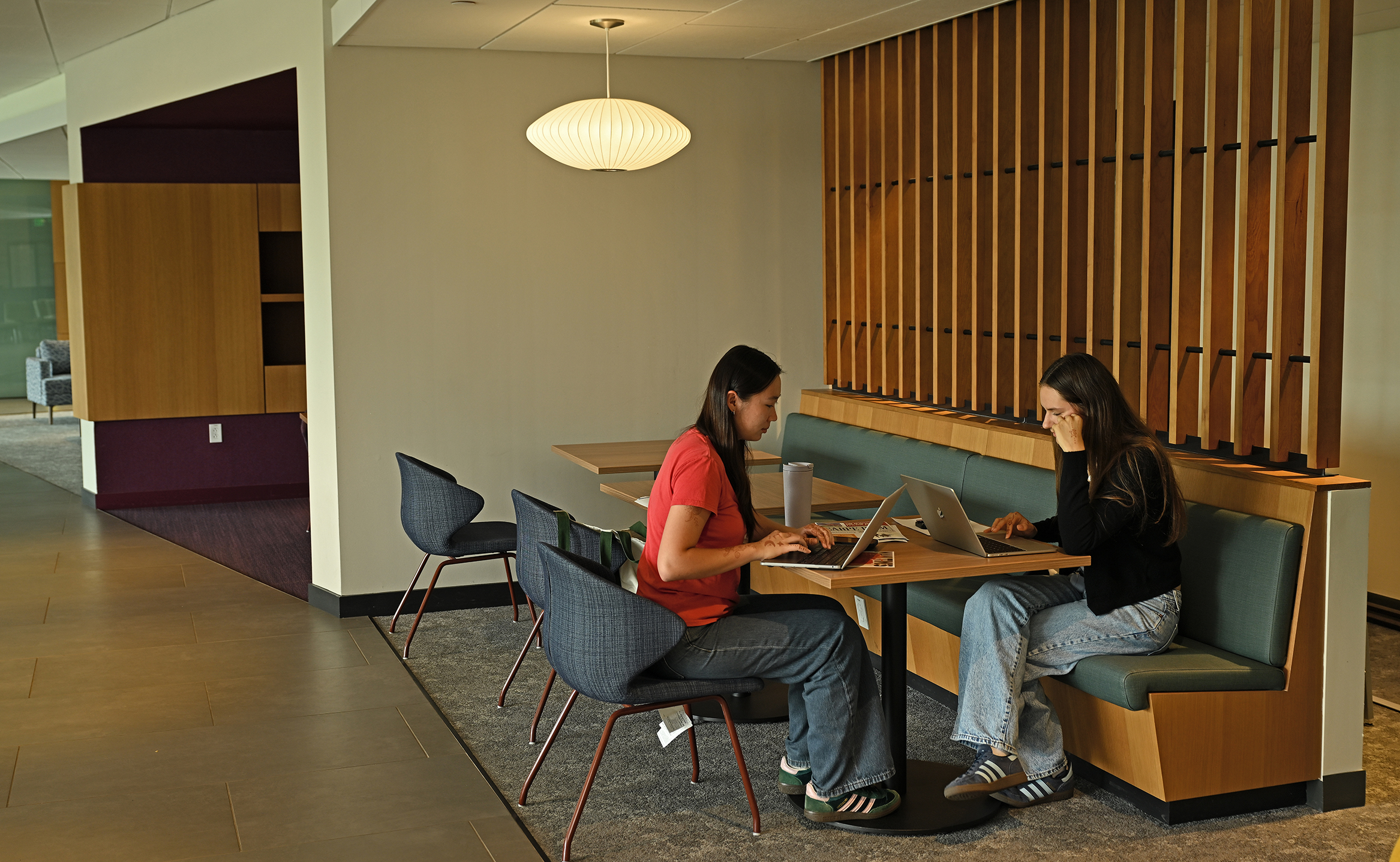 Two female college students sit at a table and work on laptop computers in a college student center.