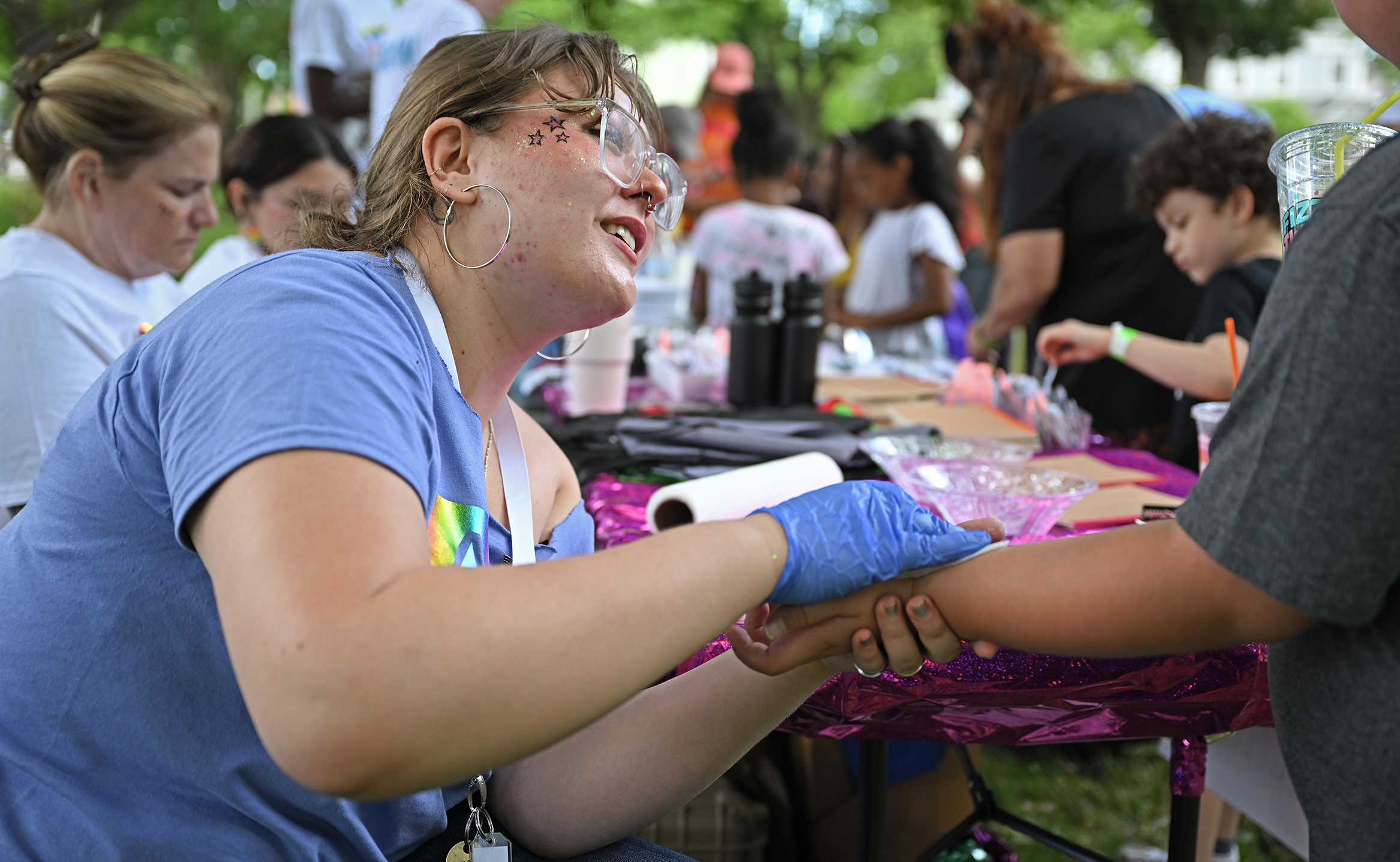 A female college students with large eyeglasses hoop earrings and temporary star tattoos on her cheeks smiles as she looks at a child who's hand she is holding while applying a temporary tattoo at a table in an outdoor setting. .