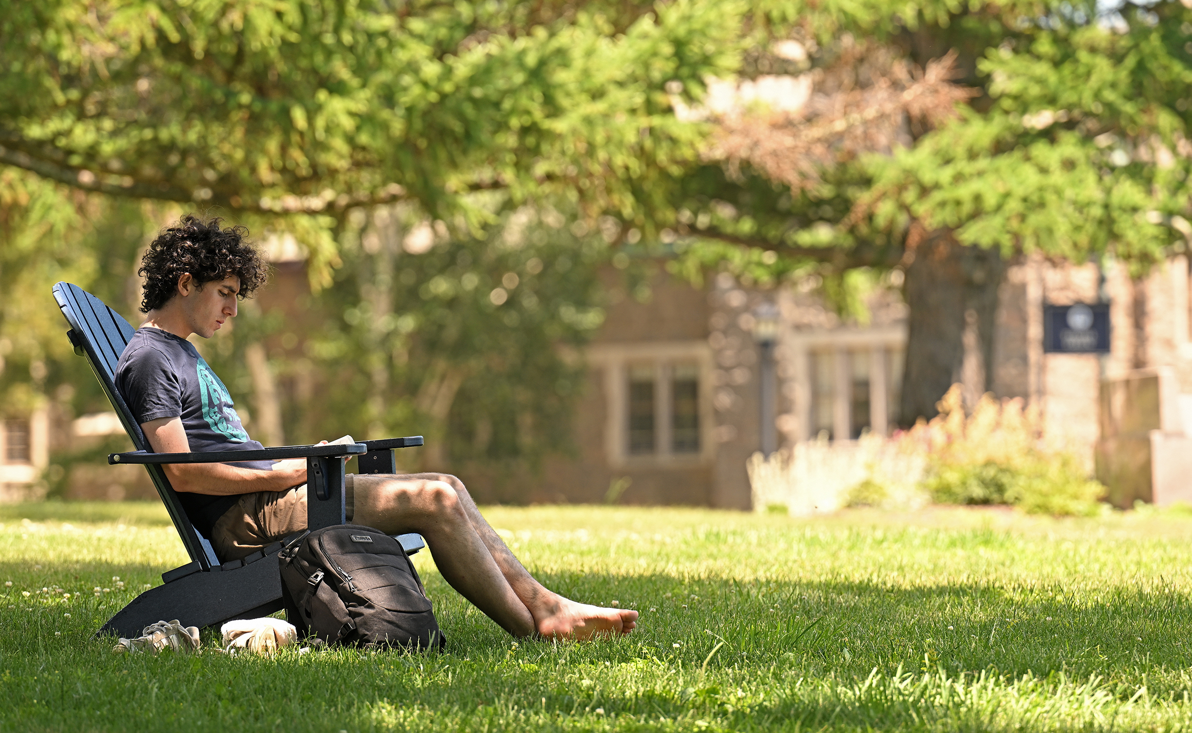 A college student in short pants and bare feet sits and reads in an Adirondack style chair in the shade of a tree on a college green with granite buildings in the distance.