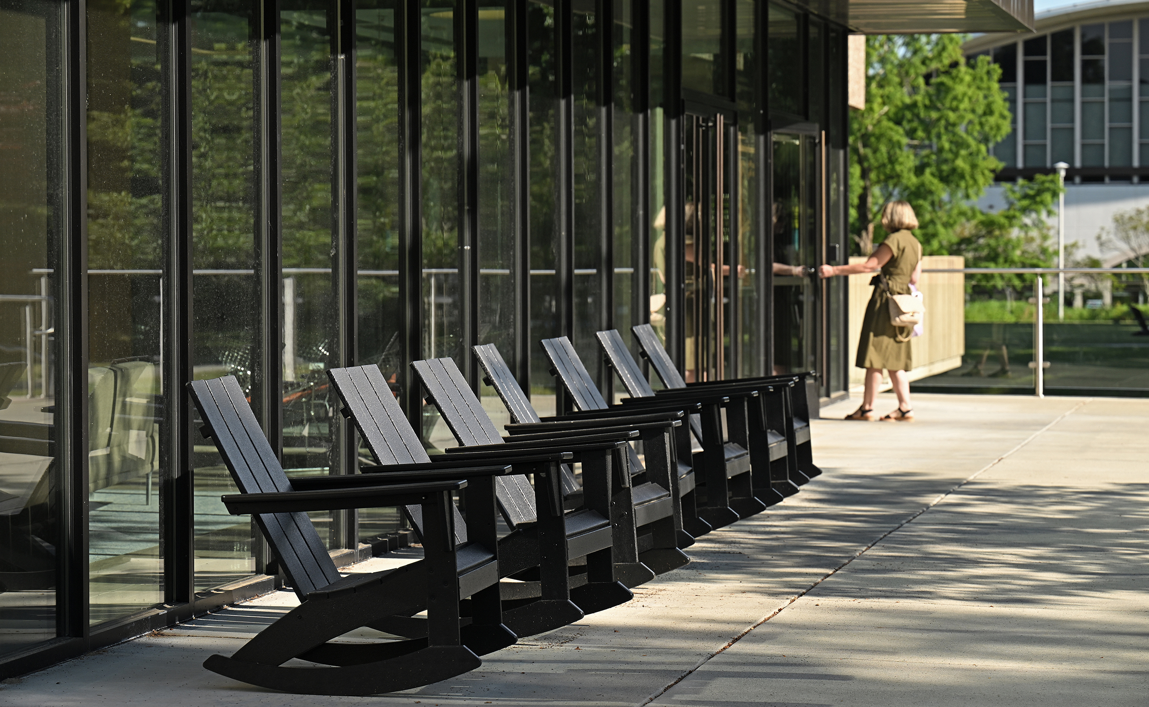 A college library employee opens the building door alongside a row of Adirondack style rocking chairs in front of a glass-front wall on a concrete patio.