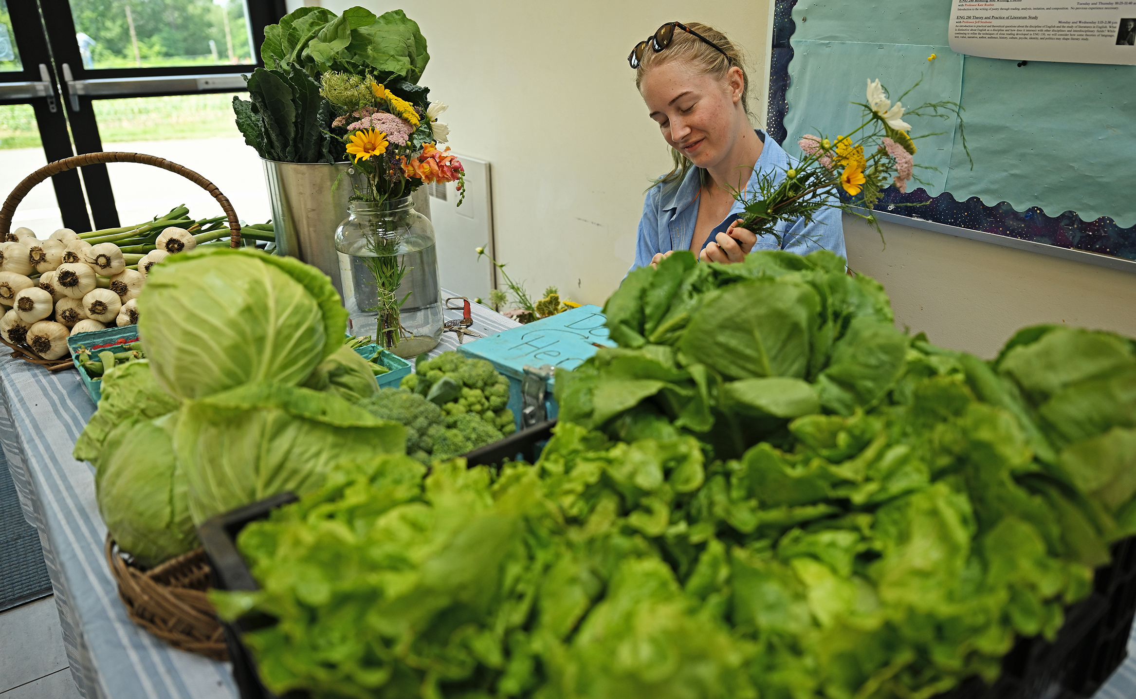 A female college students sits at a table covered in produce and assembles bouquets of wildflowers.