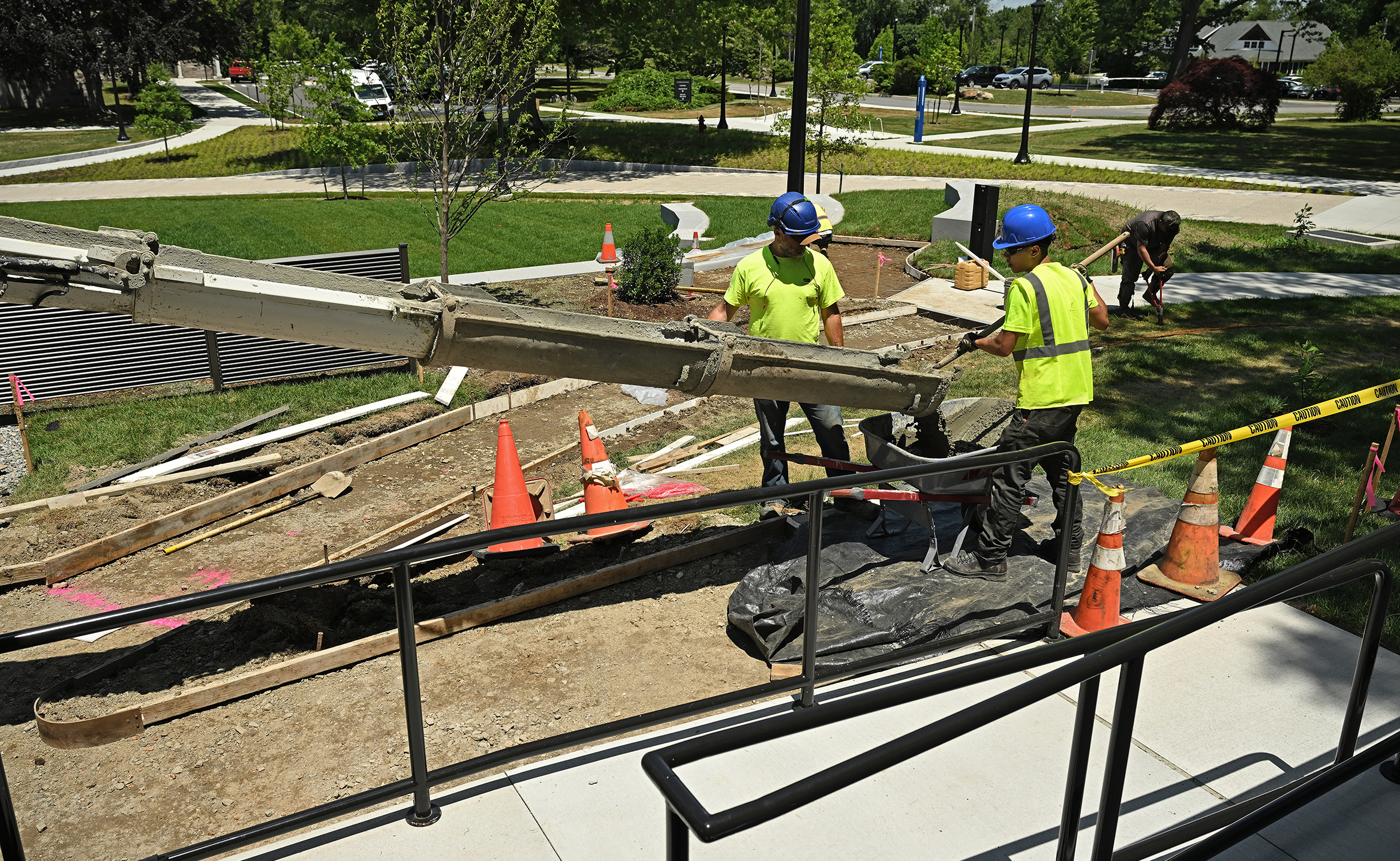 Two construction workers in bright yellow vests and blue hardhats guide a chute pouring concrete to a wheelbarrow at a walkway construction site on a college campus.