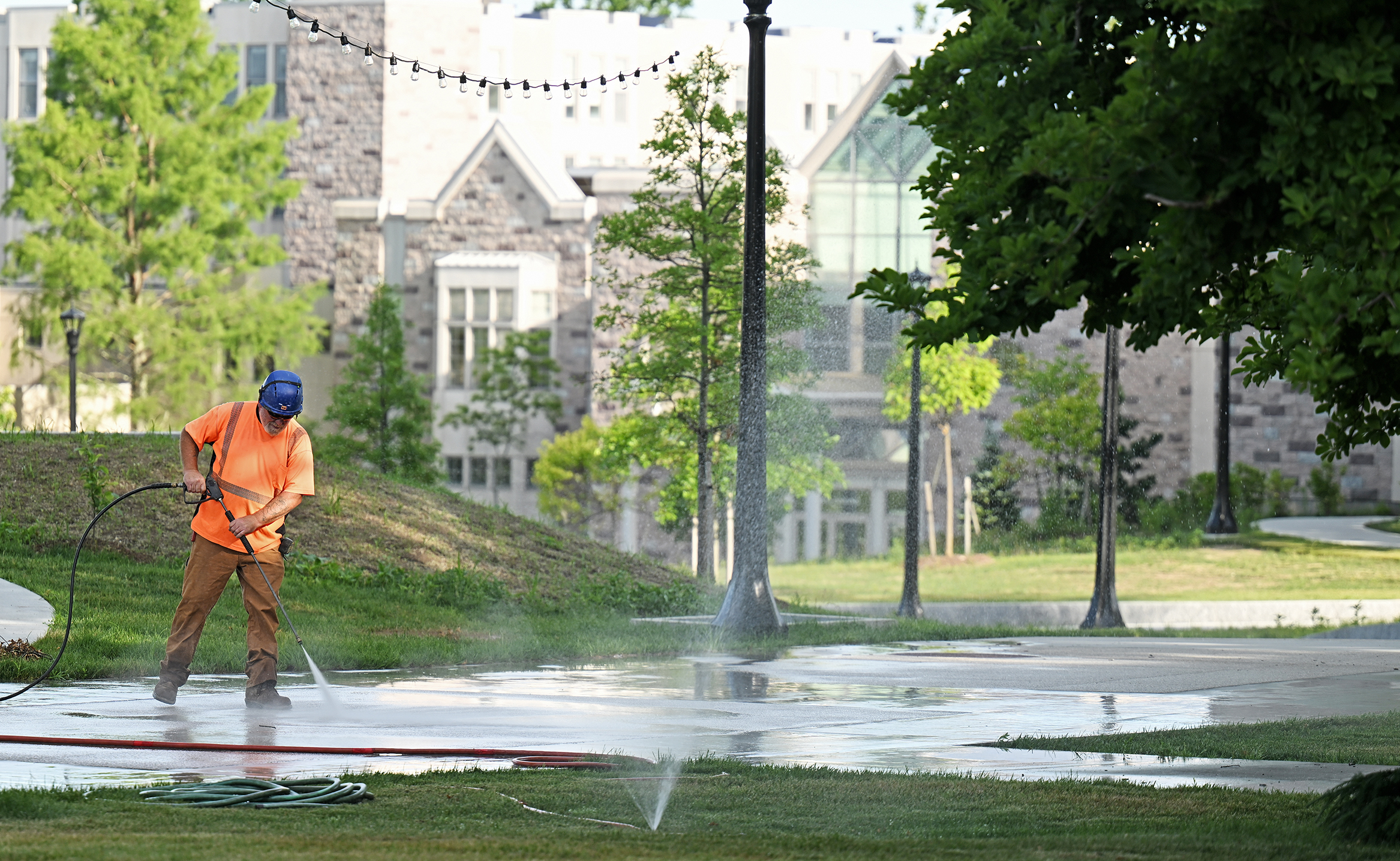 A contractor in orange shirt and blue hardhat uses a power washing wand to clean a pedestrian walkway lined with light poles and with a granite-faced college building in the distance.