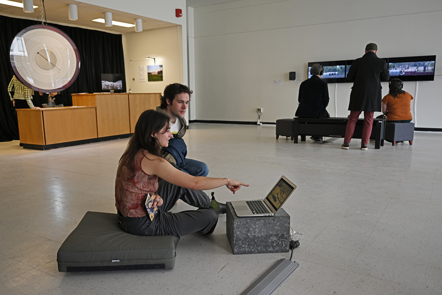 Visitors to a gallery showing art and technology works view a piece on display on a laptop computer while seated on the floor as other visitors view other pieces in the background.