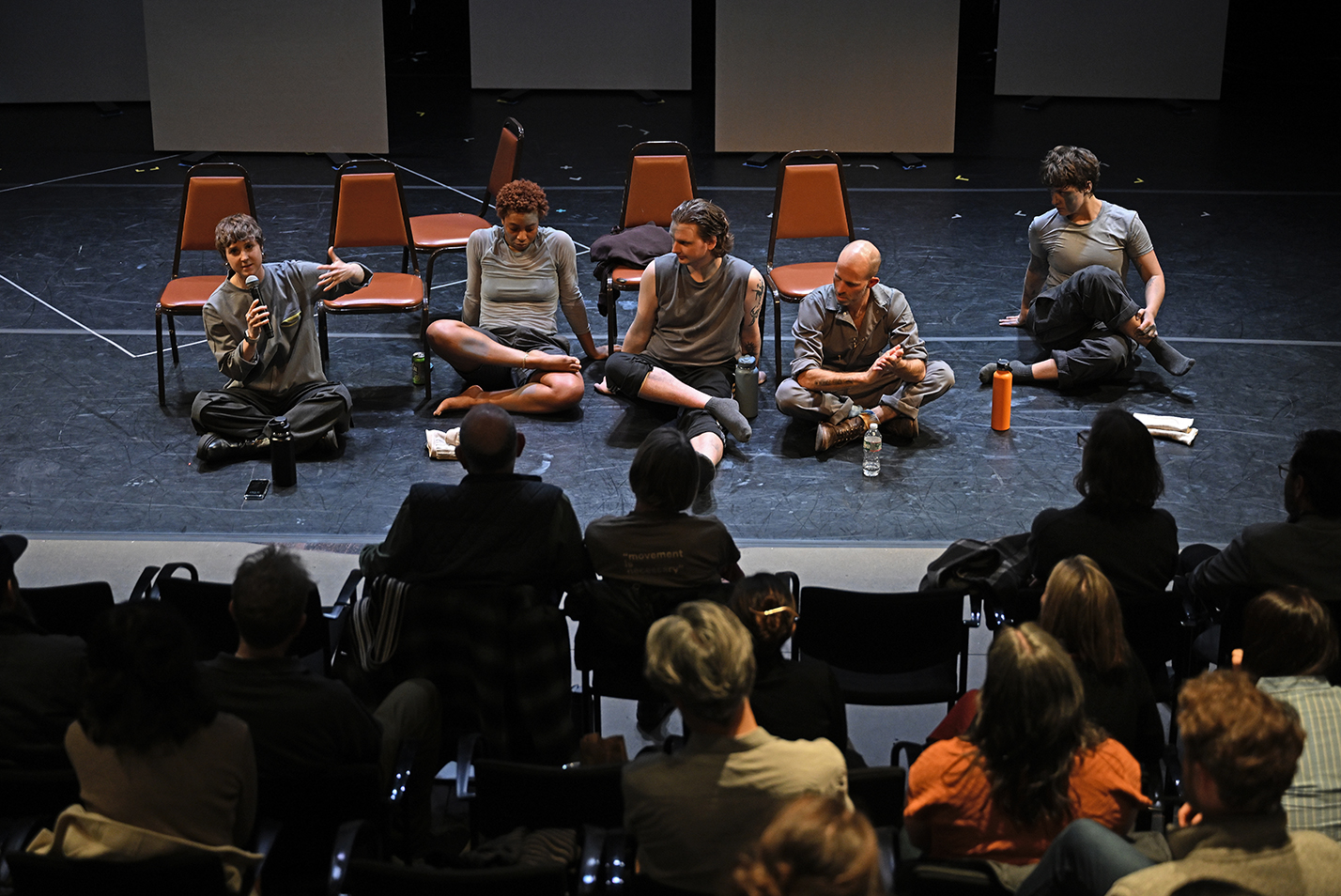 A group of dance performers and their choreographer sit on the floor of a black box theater and answer questions from the audience following a performance.