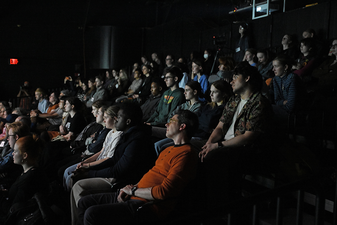 A full house audience in highly banked theater seating is lit from the glow from a performance on the stage in a black box theater space.