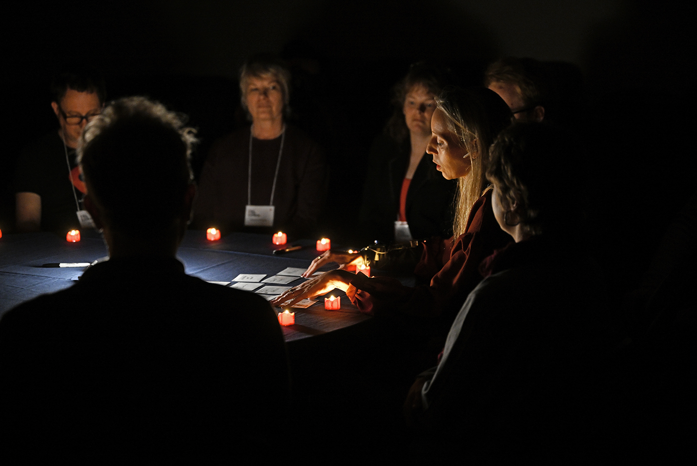 A group of people gather around a table lit with electric votive-style candles as a medium with long blond hair leads a seance in a dark room.