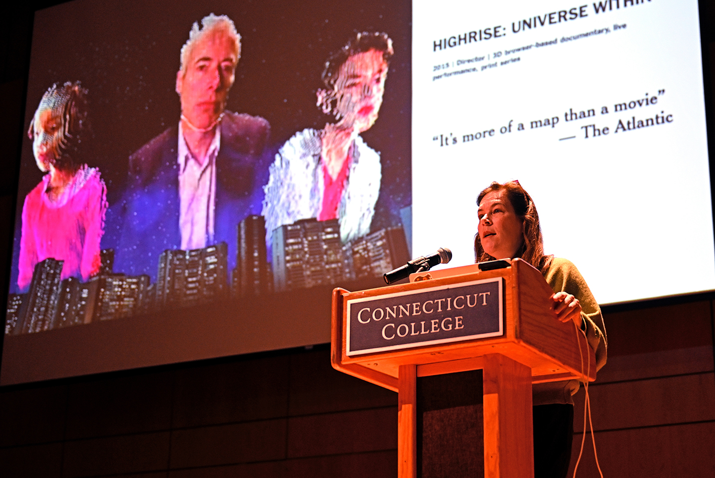 A speaker at a podium presents an address with a screen displaying three computer generated people behind her.